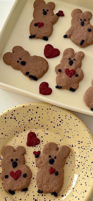 Galletas en forma de quokka con corazones rojos sobre una bandeja color crema y un plato amarillo con motas negras.