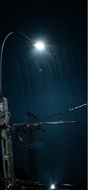 暗い夜空の下、激しい雨を照らす明るい街灯と電線のシルエット。