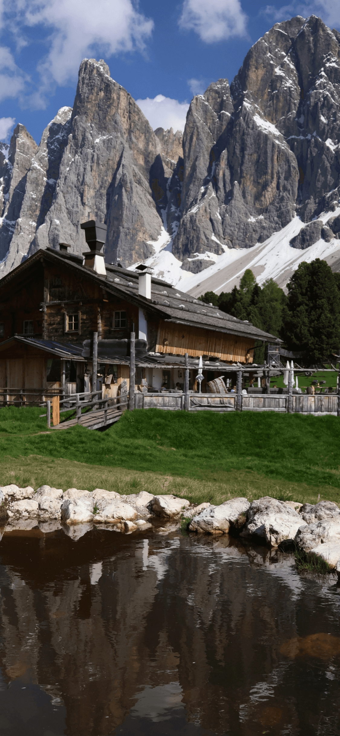 多洛米蒂山脈下的傳統木結構小屋，背景是險峻的山峰，前方有池塘倒影。