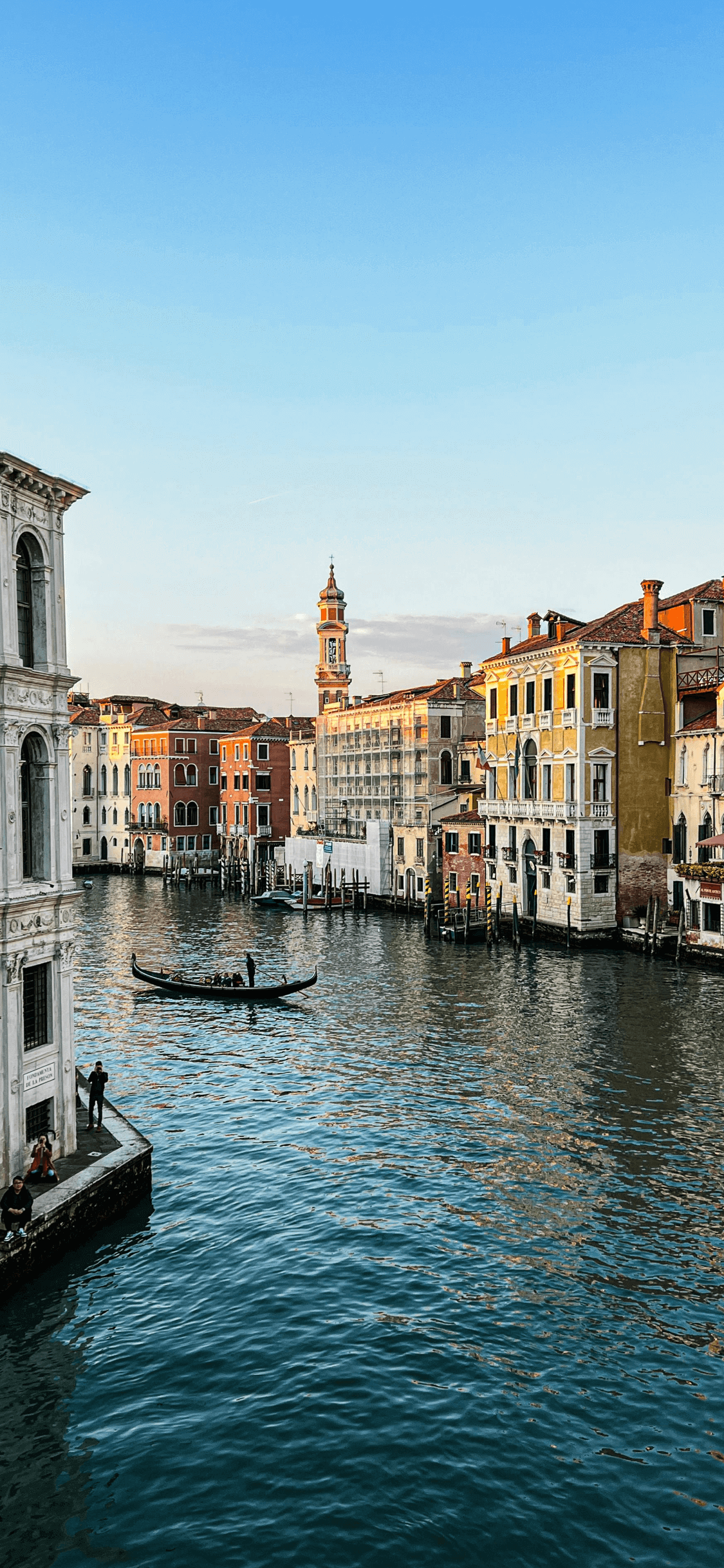 威尼斯大運河的垂直構圖攝影，畫面中有貢多拉船、橙黃色的歷史建築以及夕陽下的晴朗藍天。